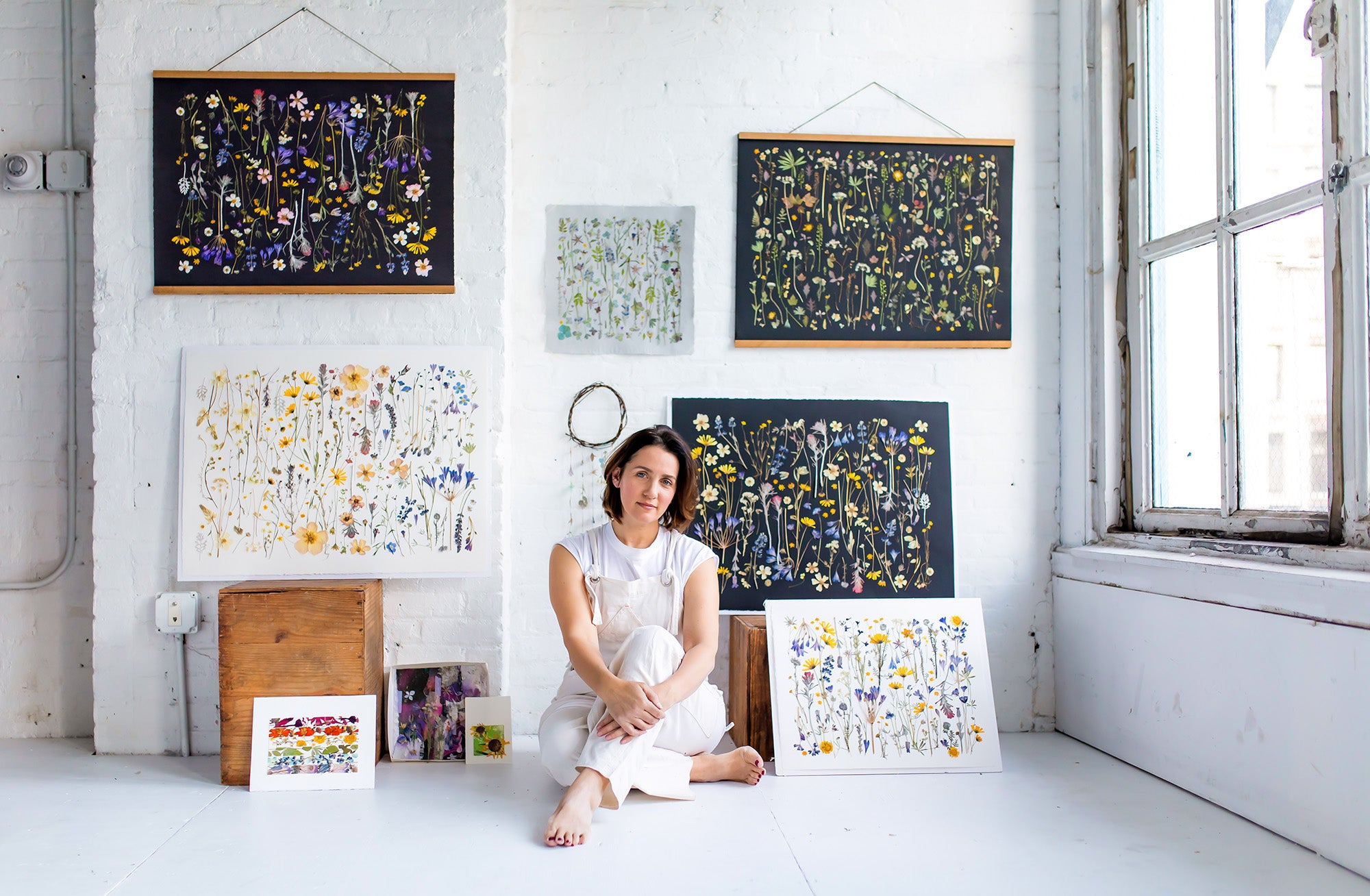Woman sitting on the floor surrounded by framed artworks in a bright room.