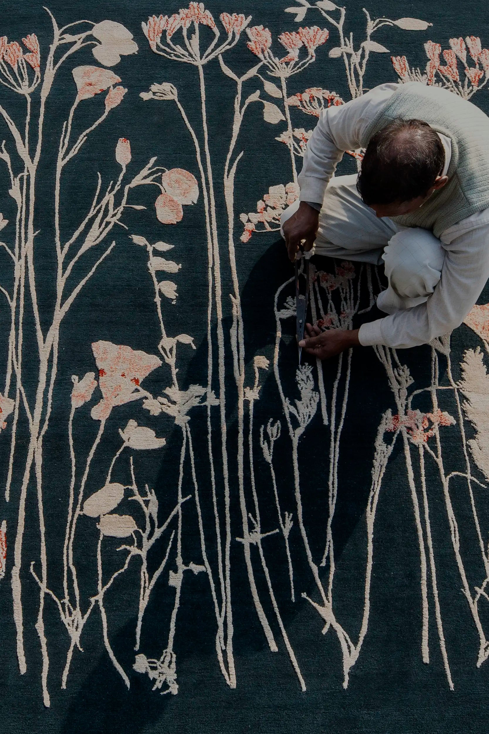 A person crouches, meticulously trimming a carpet with floral patterns on a dark background. The scene exudes craftsmanship and focus.