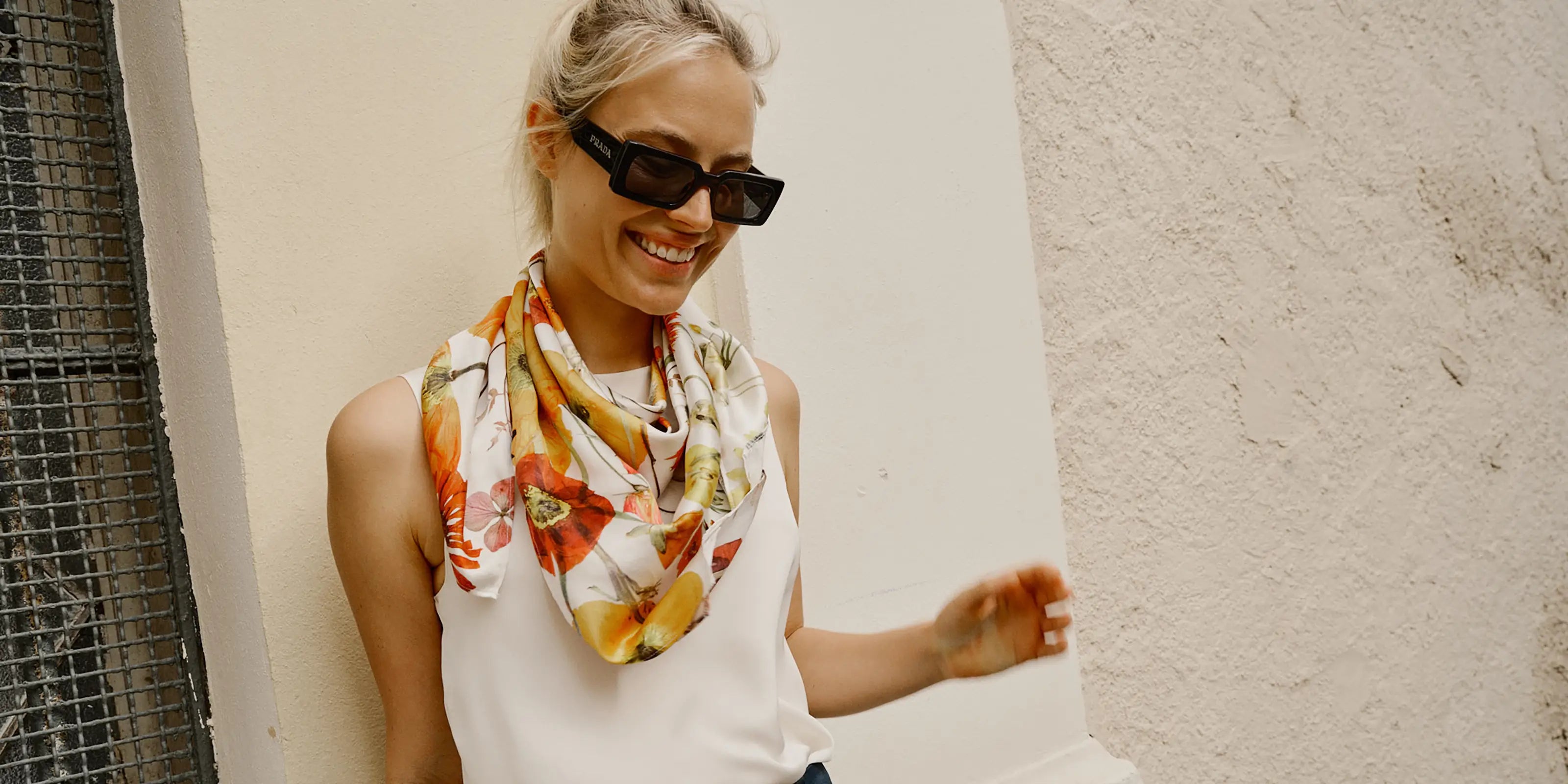 Woman in sunglasses smiling, wearing a floral scarf and white top, stands against a textured wall. Bright and cheerful, exuding a casual vibe.