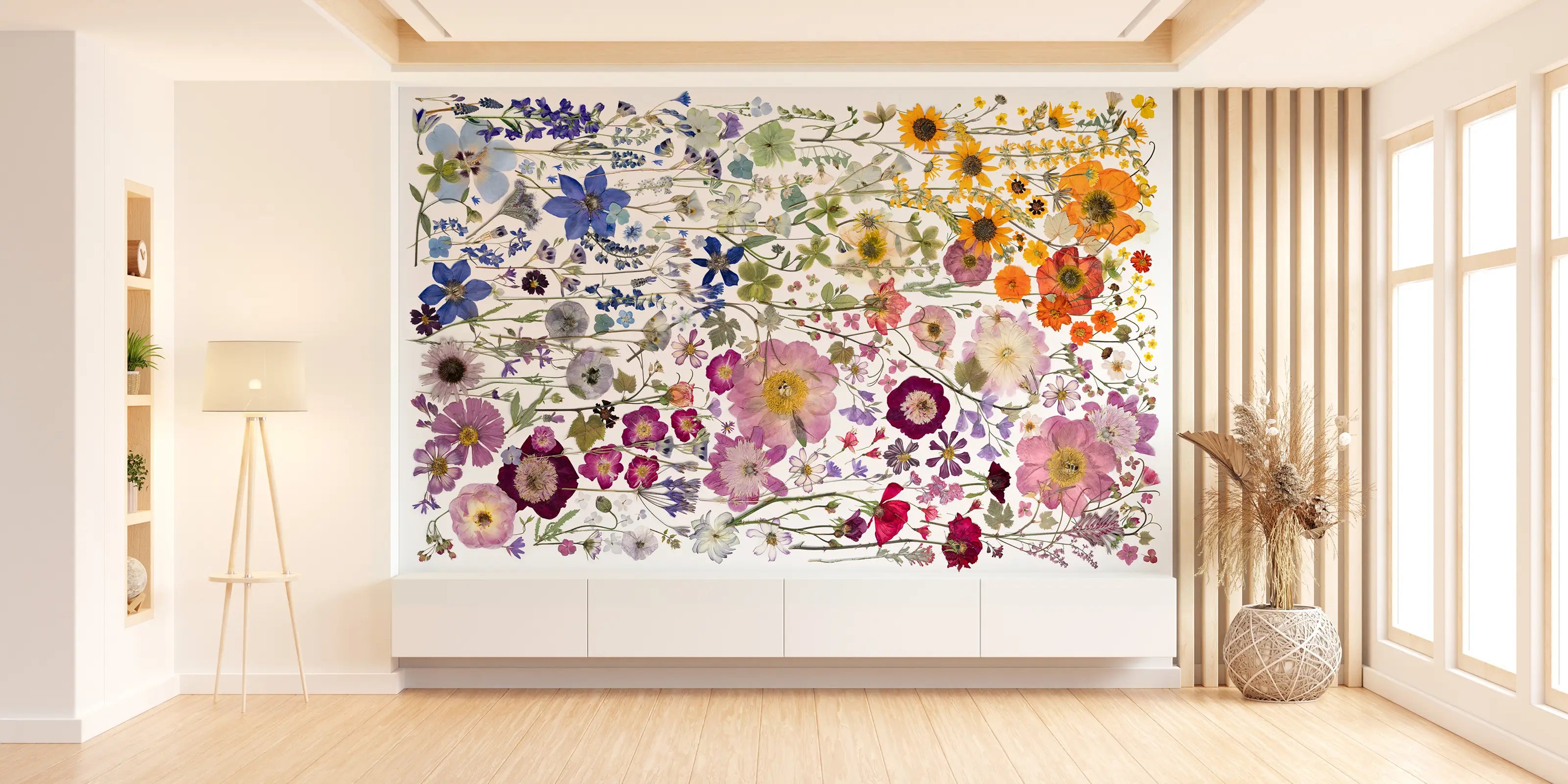 A bright, modern living room features a large, colorful floral mural on the back wall. The mural displays various flowers in blues, purples, yellows, and oranges, creating a lively and vibrant focal point. A minimalist lamp and decorative vase with pampas grass complement the serene space.