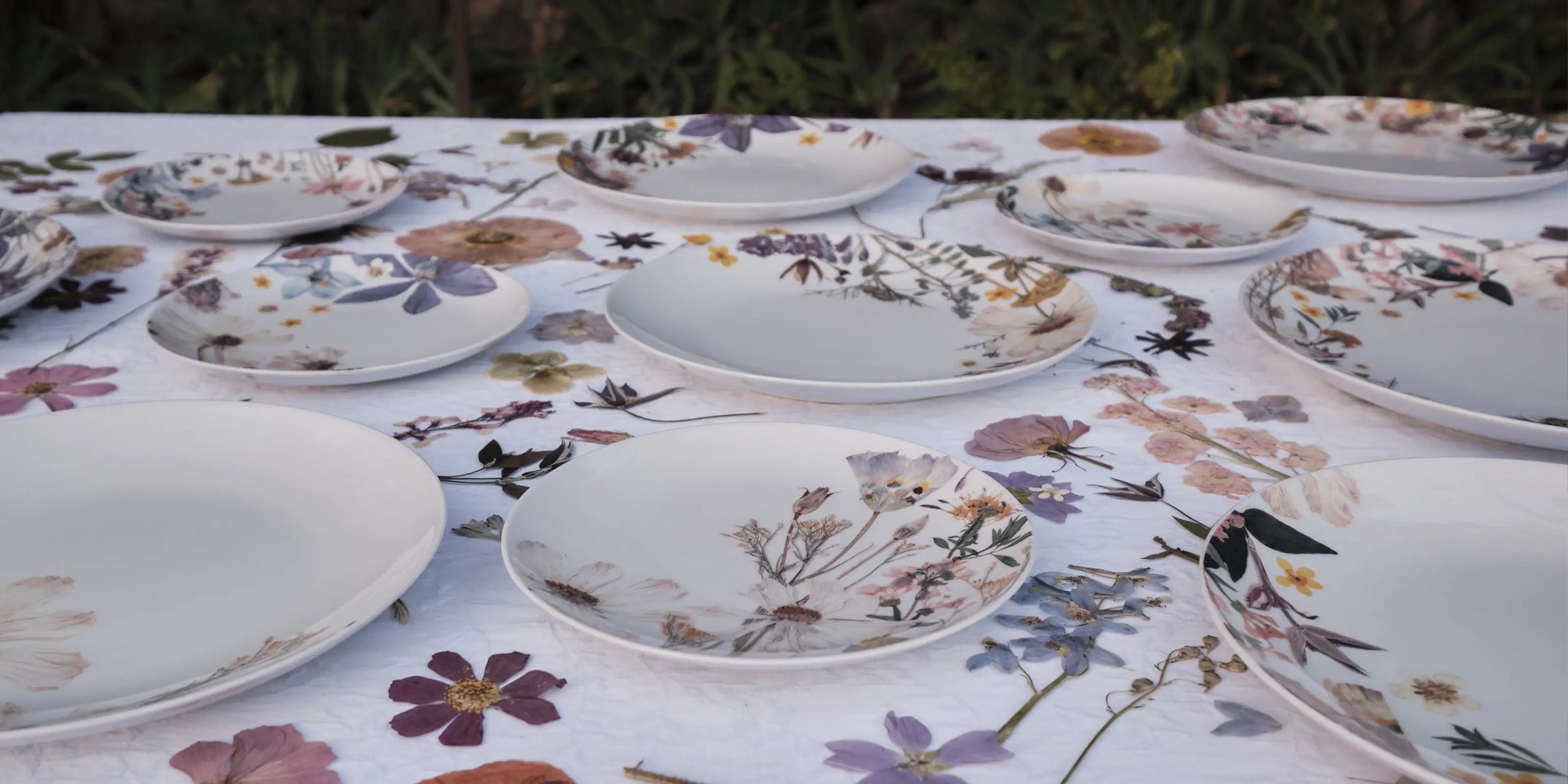 A table is set with white plates adorned with floral designs, matching the tablecloth's scattered pressed flowers. The scene evokes a serene, elegant ambiance.