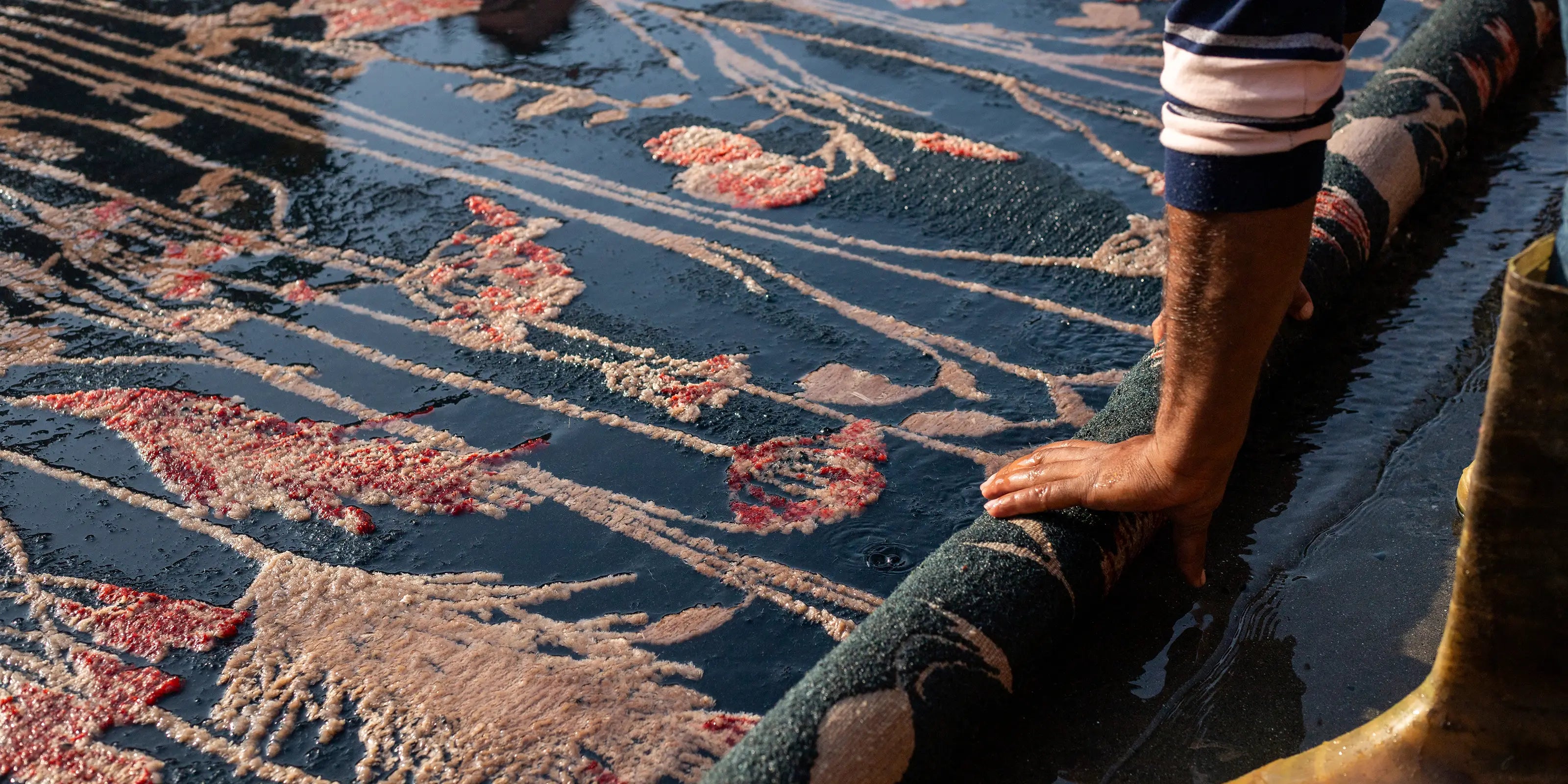 A person rolls up a wet, floral-patterned carpet, with their hand emphasizing texture and labor. Sunlight highlights the vivid colors and soaked fabric.