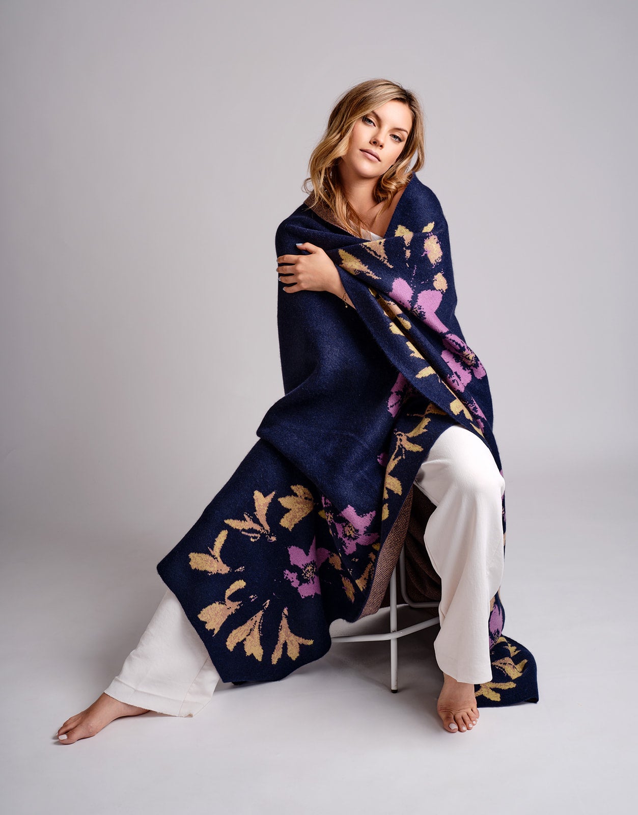 Woman sitting on a chair wearing a dark blue shawl with floral patterns on a plain background